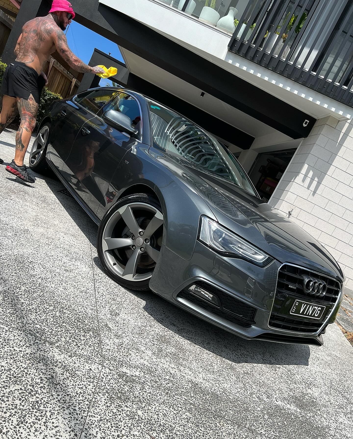 Man wearing pink headgear and black shorts washing a dark grey Audi car in a driveway.