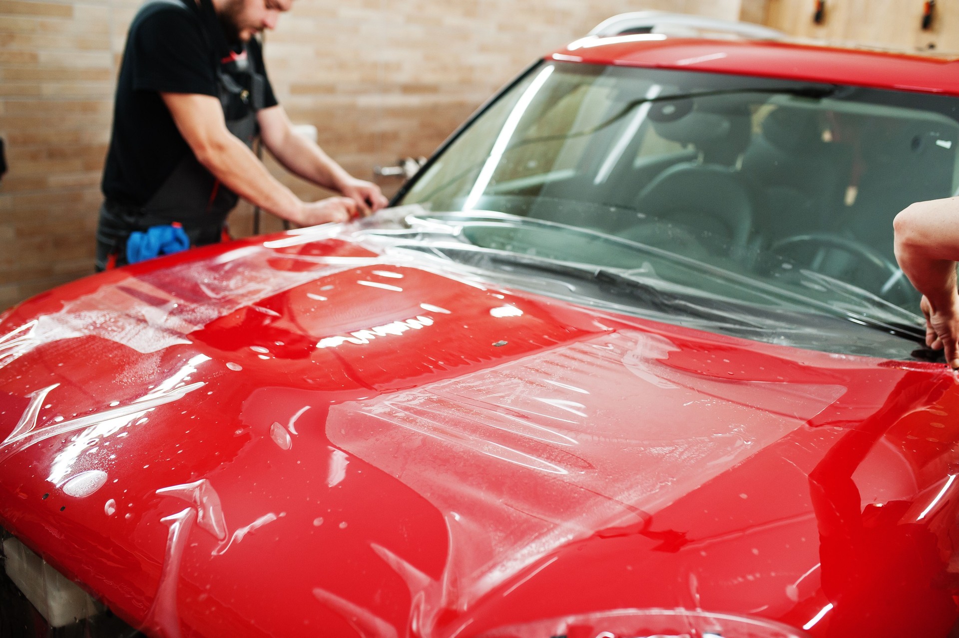 Car service worker put anti gravel film on a red car body at the detailing vehicle workshop. Car protection with special films.